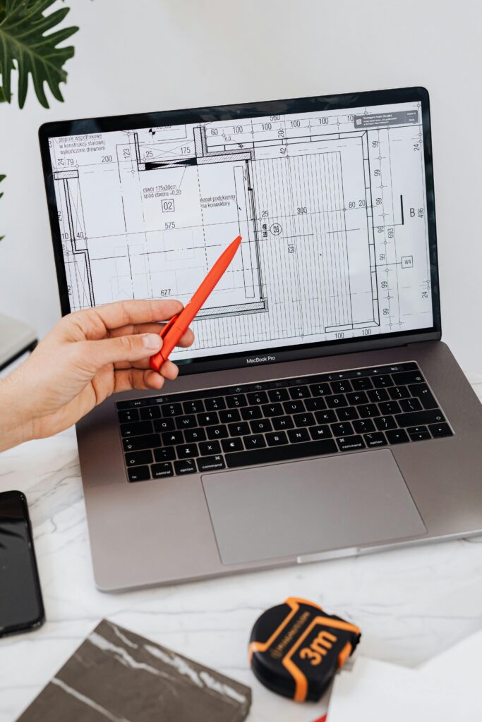 Home A hand using a red pen points at a floor plan displayed on a laptop, showcasing architectural design.
